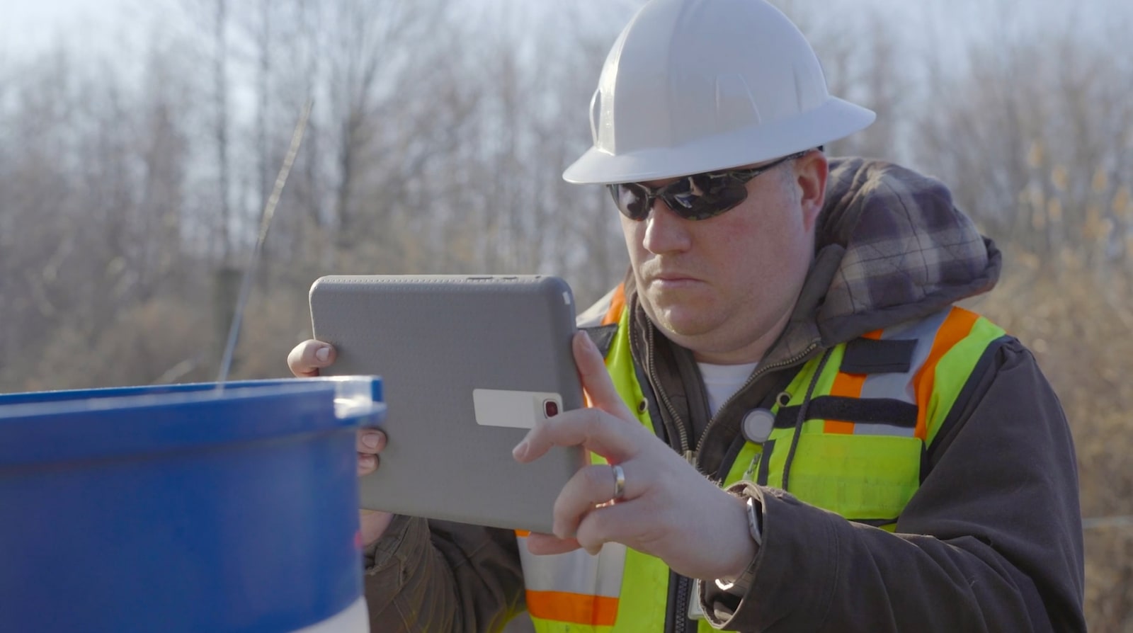 man wearing a hard hat and high visibility vest taking a photo using a tablet of a chemical barrel